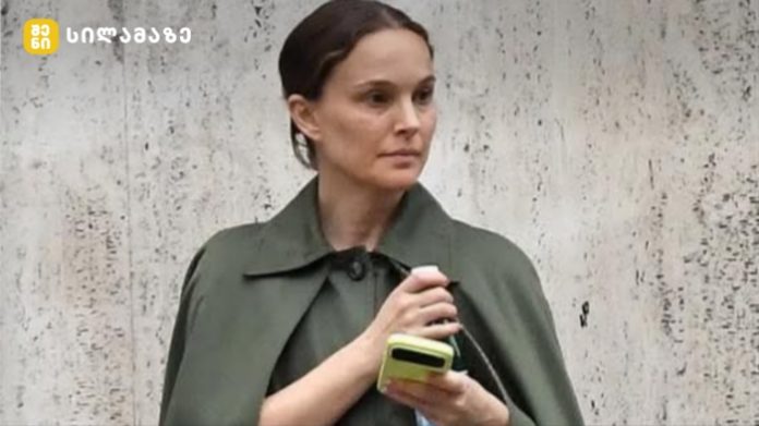 Woman with brown hair in an olive-green coat holding a lime-colored smartphone while standing against a textured concrete wall.