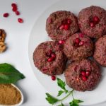 Plate of round beetroot patties topped with pomegranate seeds on a white platter with greens and spices nearby