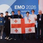 Group of young men on stage holding a Georgian flag with medals at EMO 2026 event, backdrop reads EMO 2026 April 23-29.