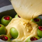 Green apples stuffed with red berries in a round cake pan, as creamy batter is poured over them.