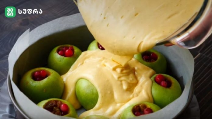 Green apples stuffed with red berries in a round cake pan, as creamy batter is poured over them.
