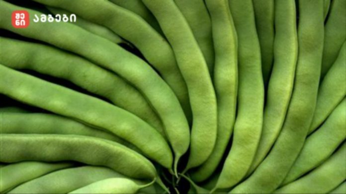Close-up of fresh green beans stacked in a dense arrangement, with a small red logo in the top-left corner.