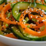 Close-up of a cucumber and carrot salad in a white bowl, topped with white and black sesame seeds.