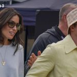 Two stylish adults wearing sunglasses walk outdoors at an event; woman in pale blue top with a heart necklace, man in a pale yellow jacket and bucket hat.