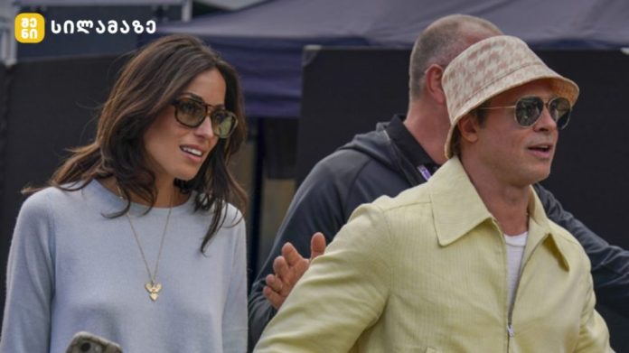 Two stylish adults wearing sunglasses walk outdoors at an event; woman in pale blue top with a heart necklace, man in a pale yellow jacket and bucket hat.