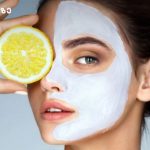 Woman with a white facial mask holding a lemon slice over her eye for skincare