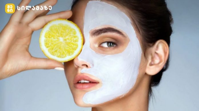 Woman with a white facial mask holding a lemon slice over her eye for skincare