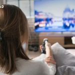 Back view of a woman sitting on a sofa, holding a remote and watching a blurred TV screen in a cozy living room.