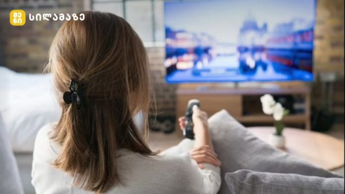 Back view of a woman sitting on a sofa, holding a remote and watching a blurred TV screen in a cozy living room.