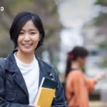 Smiling young woman with short black hair holding notebooks on a campus walkway, jacketed in denim.