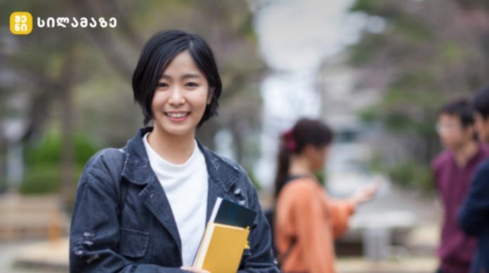 Smiling young woman with short black hair holding notebooks on a campus walkway, jacketed in denim.