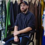 Bearded man wearing a navy hoodie and cap sits on a chair in a clothing studio surrounded by colorful garments on racks behind him.