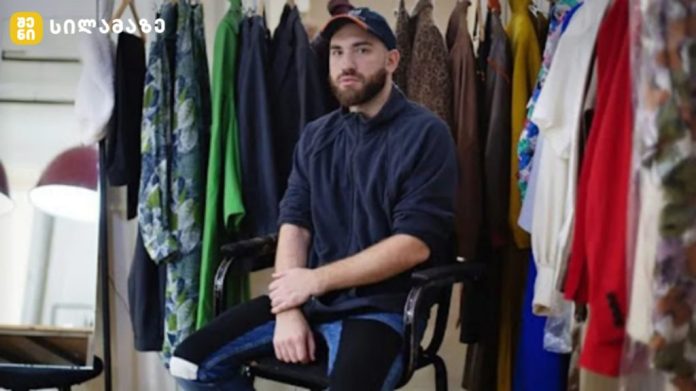 Bearded man wearing a navy hoodie and cap sits on a chair in a clothing studio surrounded by colorful garments on racks behind him.