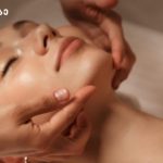 Close-up of a woman receiving a facial massage, eyes closed, as a therapist gently supports her jaw and cheekbones