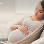 Pregnant woman relaxing on sofa, cradling her baby bump and smiling at home.