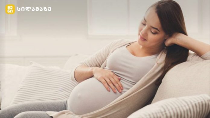 Pregnant woman relaxing on sofa, cradling her baby bump and smiling at home.