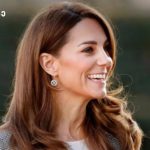 Smiling woman with long brown hair outdoors, wearing earrings, facing right side of frame with a blurred natural background.