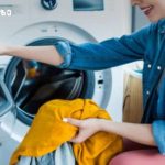 Young woman loading orange clothing into a front-loading washing machine in a laundry room.