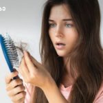 Woman with long brown hair holds a blue hairbrush and inspects strands of hair in her hand, looking surprised or worried.