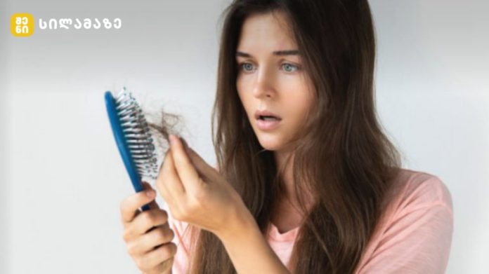 Woman with long brown hair holds a blue hairbrush and inspects strands of hair in her hand, looking surprised or worried.