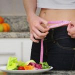 Person in a kitchen measures their waist with a pink tape measure beside a salad plate and glass of orange juice.