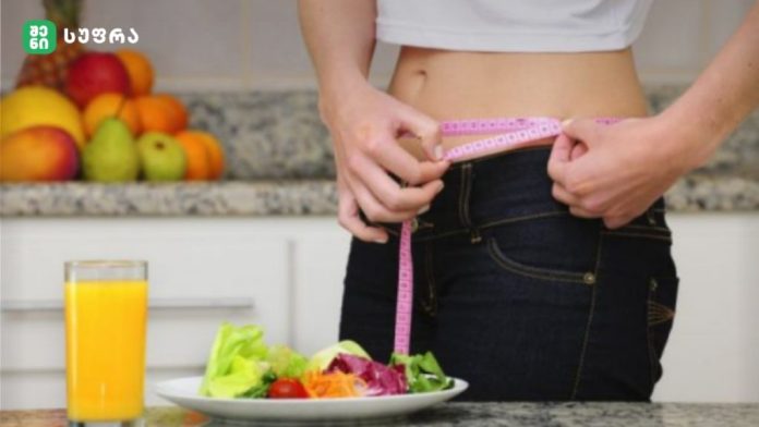 Person in a kitchen measures their waist with a pink tape measure beside a salad plate and glass of orange juice.