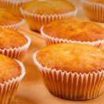 Golden muffins in white paper cups resting on a wooden countertop, fresh and warm.