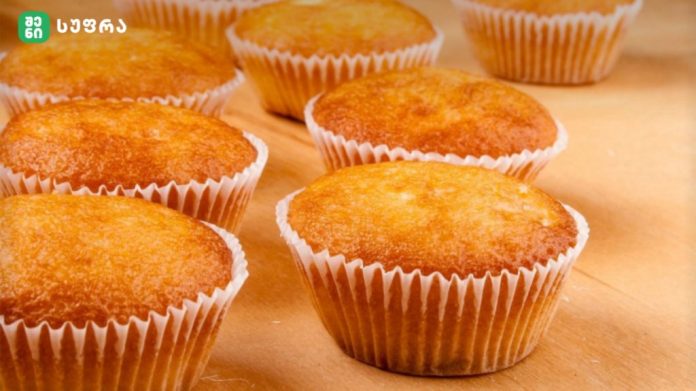 Golden muffins in white paper cups resting on a wooden countertop, fresh and warm.