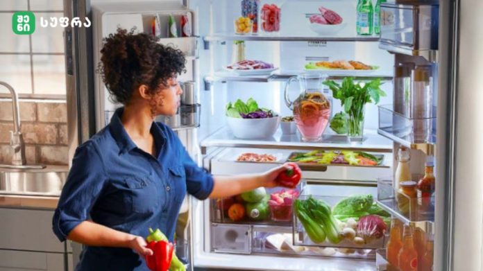 Woman in a blue shirt stands by an open refrigerator, reaching for a red pepper among fresh vegetables and drinks inside the fridge.
