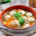 Bowl of meatball soup with carrots and fresh basil, served with bread on a light tablecloth, in a rustic pot.