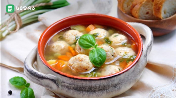 Bowl of meatball soup with carrots and fresh basil, served with bread on a light tablecloth, in a rustic pot.