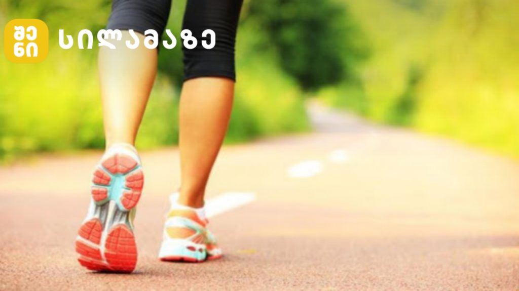Person's legs in running shoes walking on a sunlit park path; logo appears in the top-left corner.