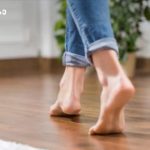 Barefoot person walking on a wooden floor indoors, jeans cuffs rolled up.