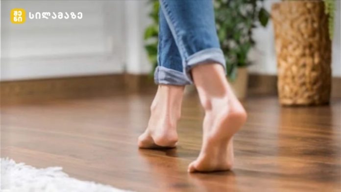 Barefoot person walking on a wooden floor indoors, jeans cuffs rolled up.