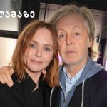 Two adults posing indoors, a woman with red hair and a man with light hair, both looking at the camera; yellow logo in the top-left corner.