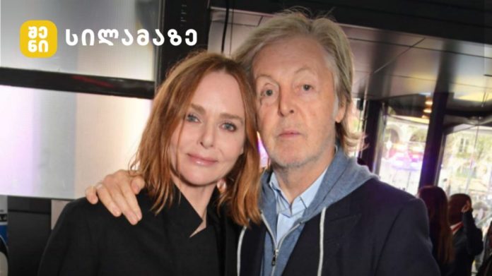 Two adults posing indoors, a woman with red hair and a man with light hair, both looking at the camera; yellow logo in the top-left corner.