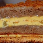 Layered cake with yellow custard filling and orange topping, displayed on a plate beside a white flower garnish.