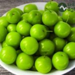 White bowl piled with bright green plums on a dark wooden surface.