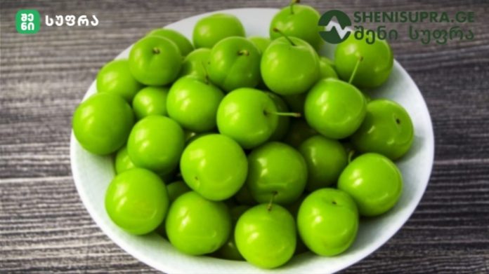 White bowl piled with bright green plums on a dark wooden surface.