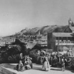 Black-and-white photo of a hillside town with a domed church and people in traditional clothing on a terrace.