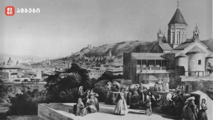 Black-and-white photo of a hillside town with a domed church and people in traditional clothing on a terrace.
