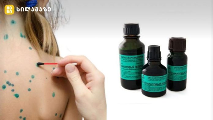 Close-up of a hand applying a green dot treat­ment on a person's back with a small brush, three dark bottles with teal labels nearby.