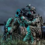 Two camouflaged soldiers kneel by a large artillery barrel in a grassy field at dusk, aiming through a scope outside the barrelled weapon