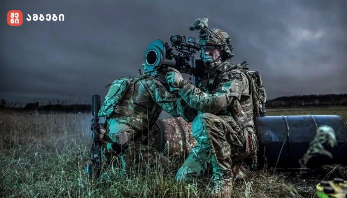 Two camouflaged soldiers kneel by a large artillery barrel in a grassy field at dusk, aiming through a scope outside the barrelled weapon