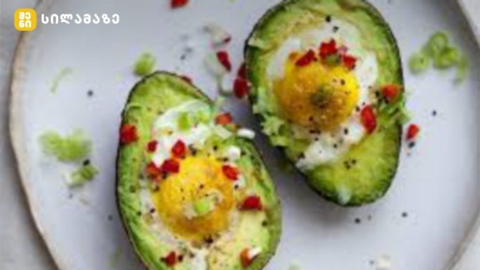 Two avocado halves hollowed and filled with boiled eggs, garnished with chopped greens and red pepper on a white plate.