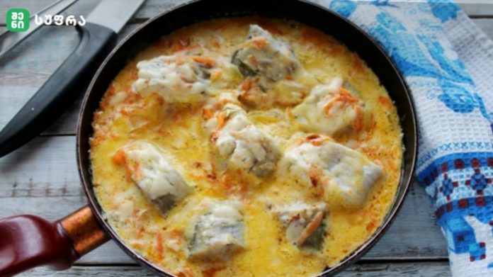 Skillet with chunks of white fish in a creamy yellow sauce with vegetables on a wooden table.