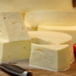 Block of soft cheese with wedges on a wooden cutting board, knife, tomatoes, garlic, and milk in the background.