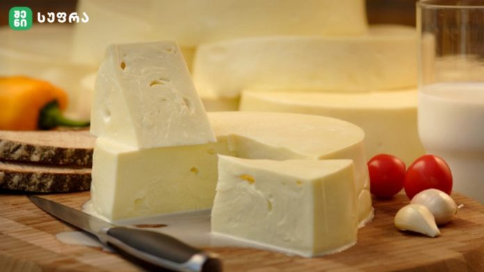 Block of soft cheese with wedges on a wooden cutting board, knife, tomatoes, garlic, and milk in the background.