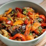 Bowl of colorful sautéed vegetables (eggplant, red and orange peppers, tomatoes) garnished with herbs on a wooden board.