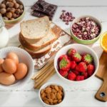 Assorted breakfast ingredients on a white wooden table: eggs, milk, sliced bread, almonds, chocolate, pistachios, strawberries, oats, and orange halves.